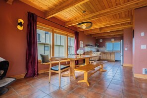 A spacious dining area with a wooden table and benches, a kitchen in the background, and large windows with curtains. - The Atlas - Sauna and Beach Access (Dr)