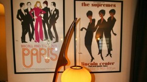 Ein Zimmer mit einem gerahmten Poster von 'Paris' und 'The Supremes' an der Wand, einer Lampe und einem roten Kissen auf einem Stuhl.