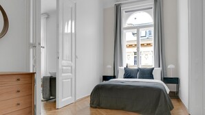 Una camera da letto con un letto, una cassettiera, una finestra con tende e vista sugli edifici esterni.