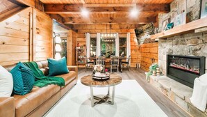 L’intérieur chaleureux d’un chalet avec un plafond en bois, une cheminée en pierre, un coin repas et un canapé agrémenté de coussins.