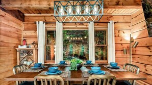 L’intérieur d’un chalet en bois présente une table à manger dressée pour six personnes, entourée de chaises, et une grande fenêtre agrémentée de rideaux.