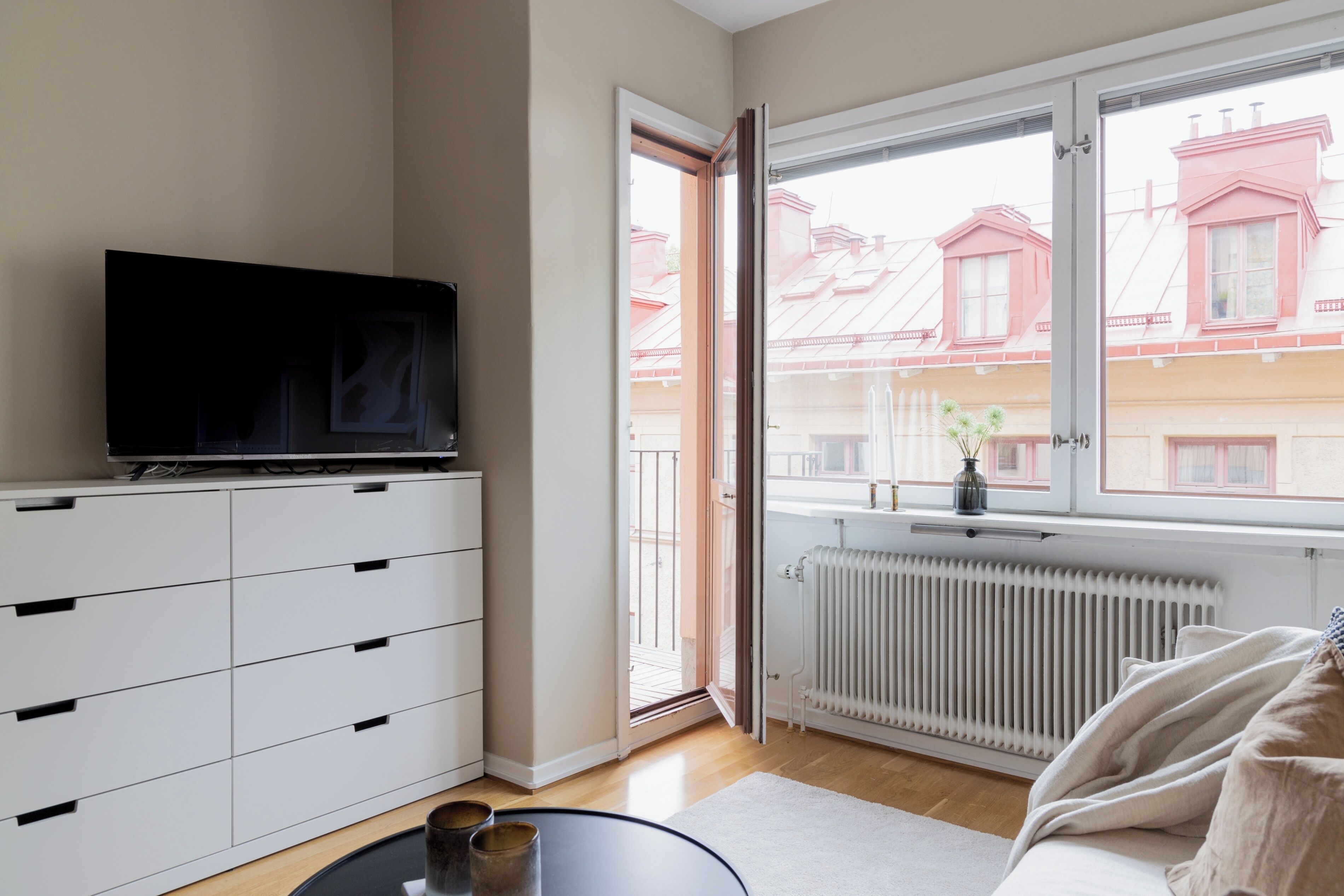 Uma sala de estar moderna com uma TV sobre um armário branco, uma janela grande com vista para edifícios vizinhos e um radiador sob a janela.
