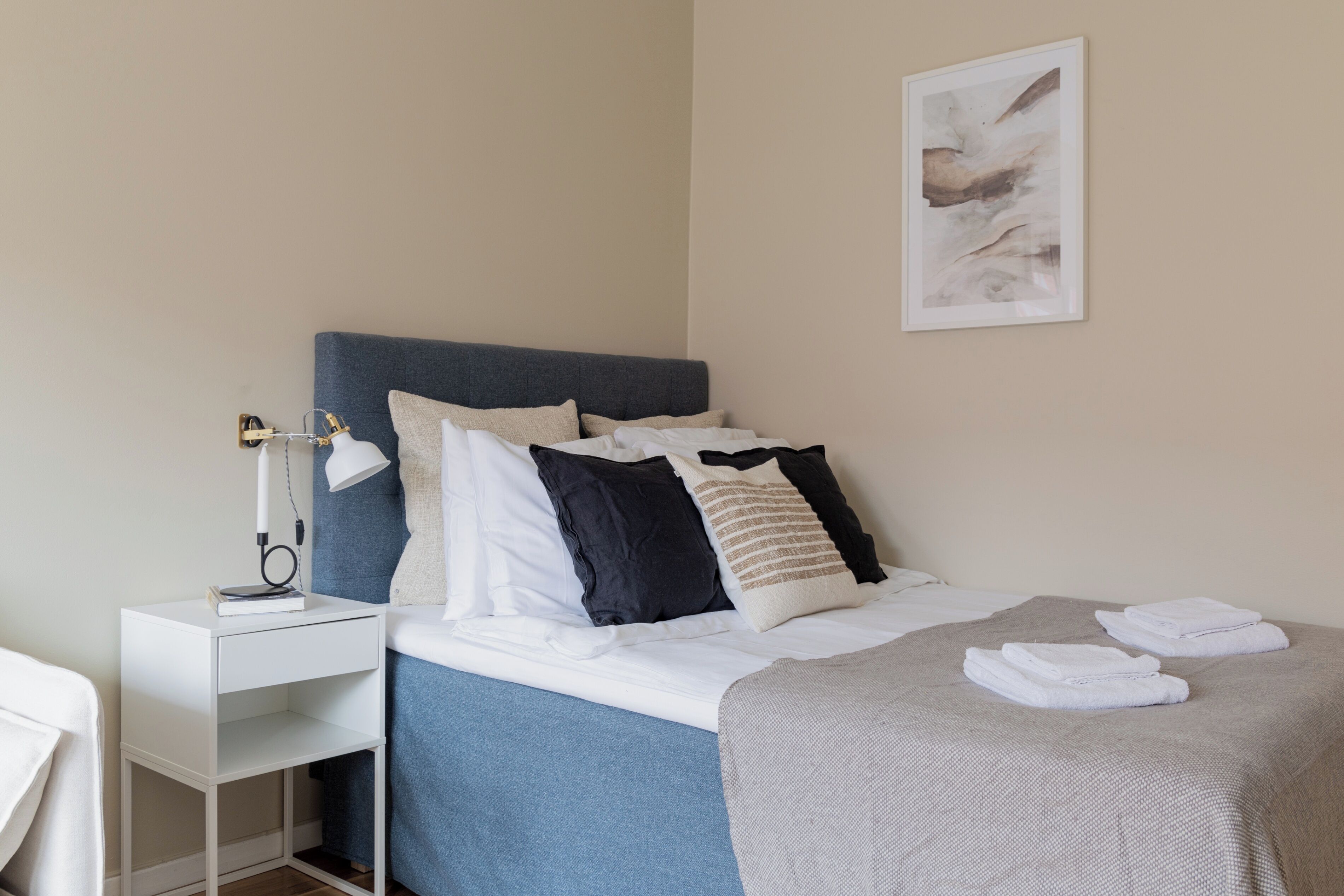 Una cama bien hecha con cabecera azul, ropa de cama blanca y una manta gris. Hay una mesita de noche con una lámpara y un cuadro en la pared.