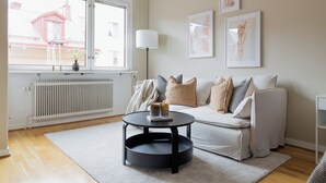 Un salon moderne avec un canapé, une table basse ronde et un radiateur. Des tableaux encadrés ornent le mur, et une fenêtre offre une vue sur les bâtiments à l’extérieur.