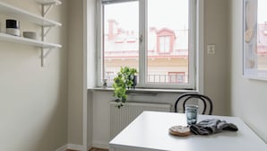 Une table de salle à manger blanche, ornée d’une plante, d’un verre d’eau et d’une serviette en tissu. Des étagères blanches, sur lesquelles sont posés des récipients, sont fixées au mur.