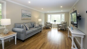 A spacious living room with a grey sofa, two armchairs, a coffee table, and a TV mounted on the wall.