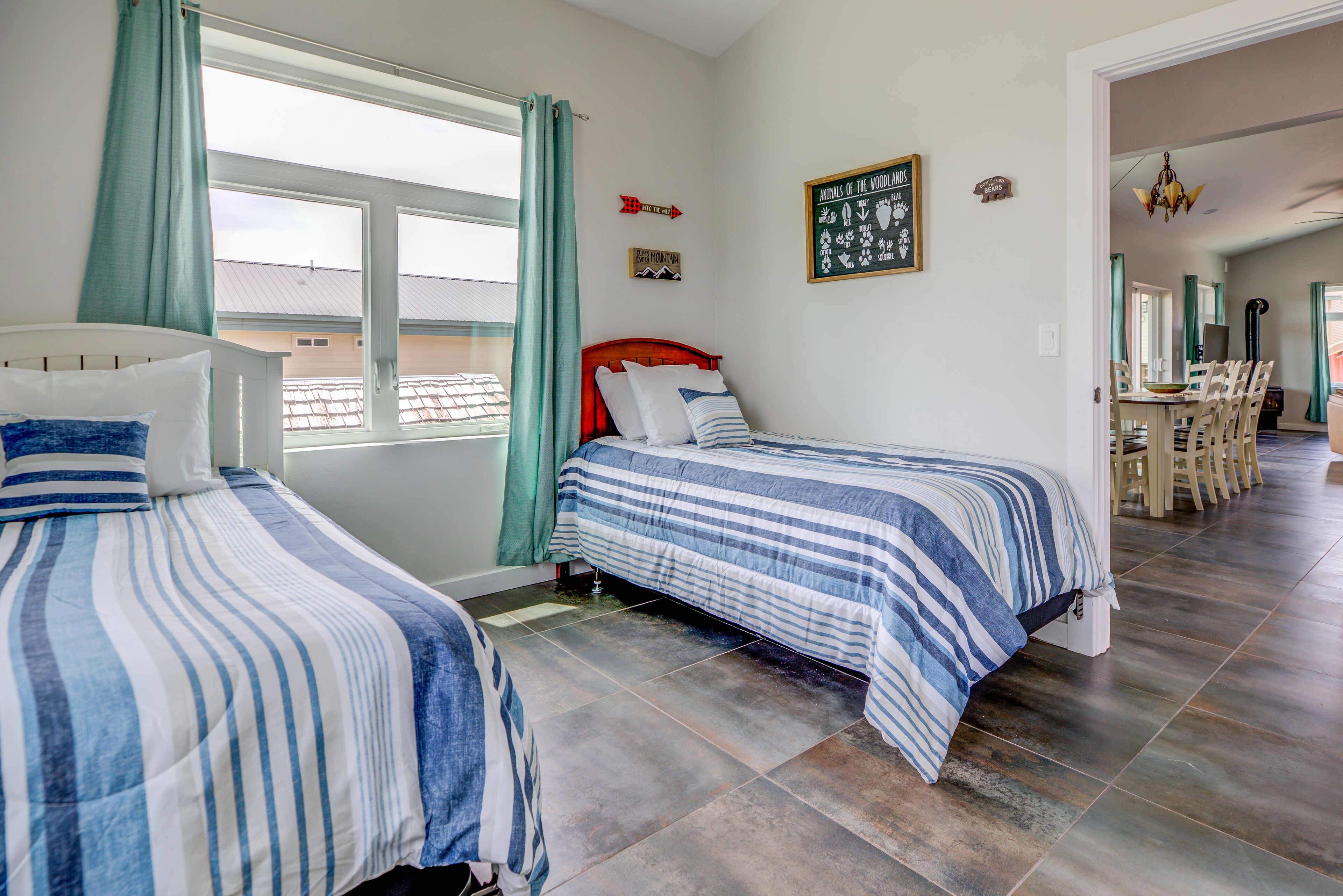 Un dormitorio con dos camas, una ventana con cortinas, una pared con un cartel enmarcado y un área de comedor visible a través de una puerta abierta.