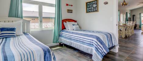Un dormitorio con dos camas, una ventana con cortinas, una pared con un cartel enmarcado y un área de comedor visible a través de una puerta abierta.