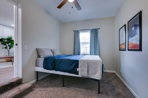 Een slaapkamer met een bed, een plafondventilator, een raam met gordijnen en een deur naar een andere kamer.