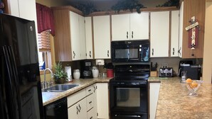 Een keuken met witte kastjes, een zwarte koelkast, een magnetron en een fornuis. Op de vloer liggen rode matten en aan de muur hangt een bord met een welkomstboodschap.