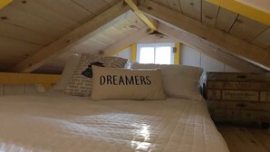 Quarto com teto inclinado, cama com colchão acolchoado, duas almofadas e uma janela.