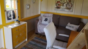 Um quarto aconchegante com teto de madeira, cômoda amarela, cadeira branca, sofá marrom e um pôster de bicicleta.