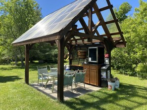 Un kiosque en bois avec un toit en métal, une table avec des chaises et un petit coin bar équipé d’un téléviseur et de placards de rangement.