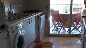 Eine kompakte Küche mit Waschmaschine, Herd und Spüle. Eine Glastür führt auf eine Außenpatio mit Tisch und Stühlen.
