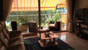 Ein Wohnzimmer mit einer Couch, einem Couchtisch und einer TV-Konsole. Ein Balkon mit Pflanzen und Blick ins Freie ist vorhanden.