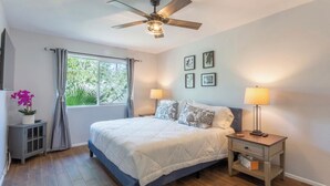 Quarto com uma cama, duas mesas de cabeceira com luminárias, um ventilador de teto, uma janela com cortinas e um pequeno armário com uma planta.