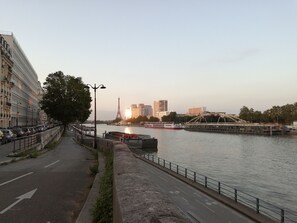 A riverside walkway with a view of the Eiffel Tower in the distance, a bridge, and modern buildings.