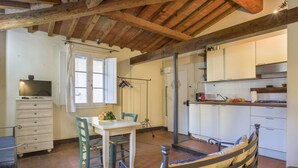 Una cocina con armarios blancos, una mesa de comedor con sillas, un televisor y una ventana con cortinas.