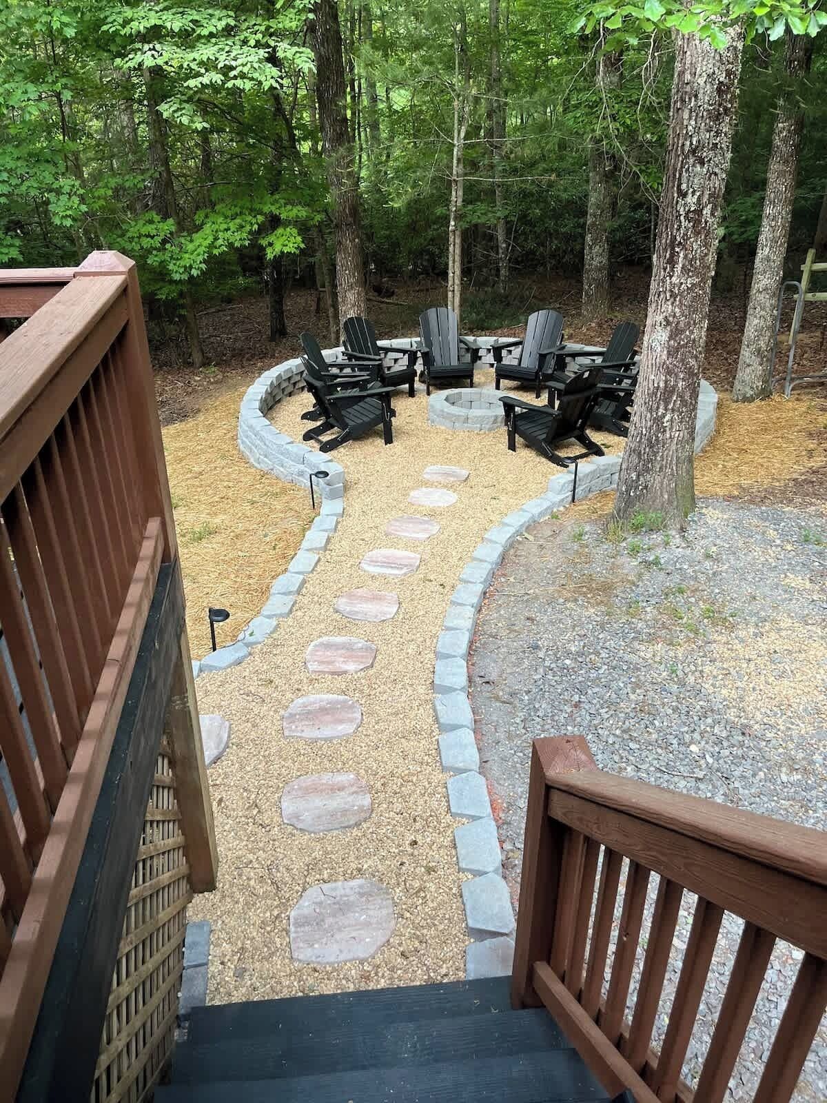 Eine Terrasse führt zu einer Feuerstelle, umgeben von Adirondack-Stühlen, mit einem Steinweg und einem Holzdeckgeländer.