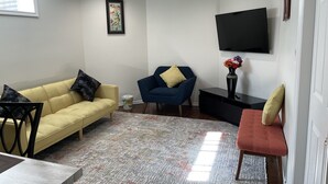 Uma sala de estar com um sofá amarelo, uma poltrona azul, um banco vermelho, uma TV preta na parede e uma obra de arte emoldurada acima do sofá.