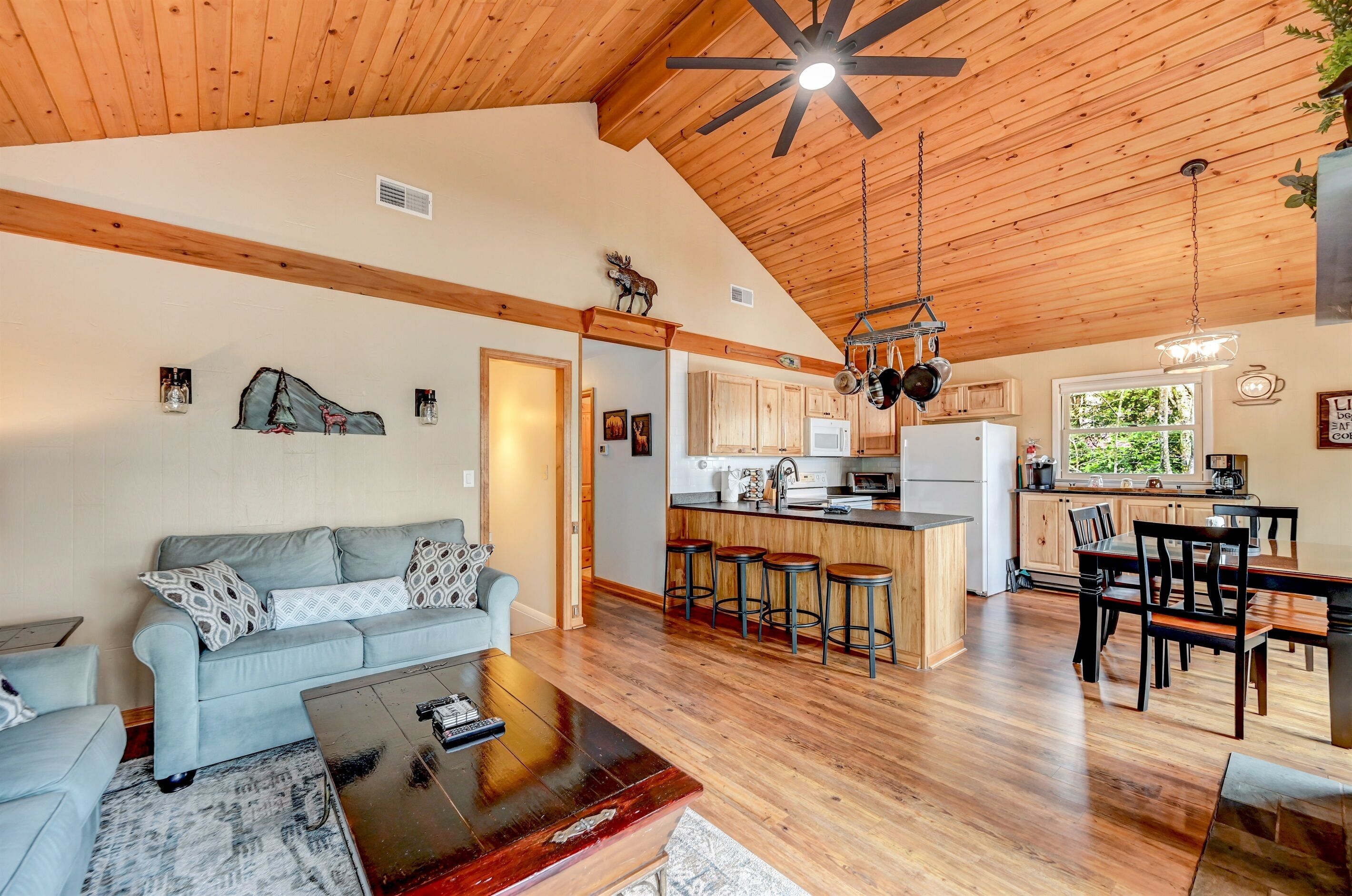 A spacious living area with a wooden ceiling, a sofa, a coffee table, and a kitchen area with a bar and stools.