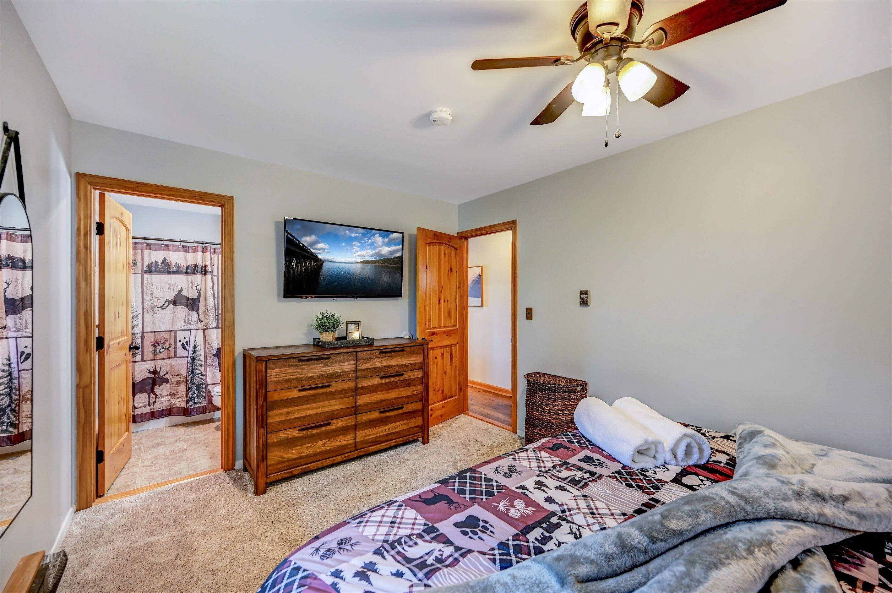 Une chambre à coucher avec un lit, une commode en bois, un ventilateur de plafond et un téléviseur fixé au mur.