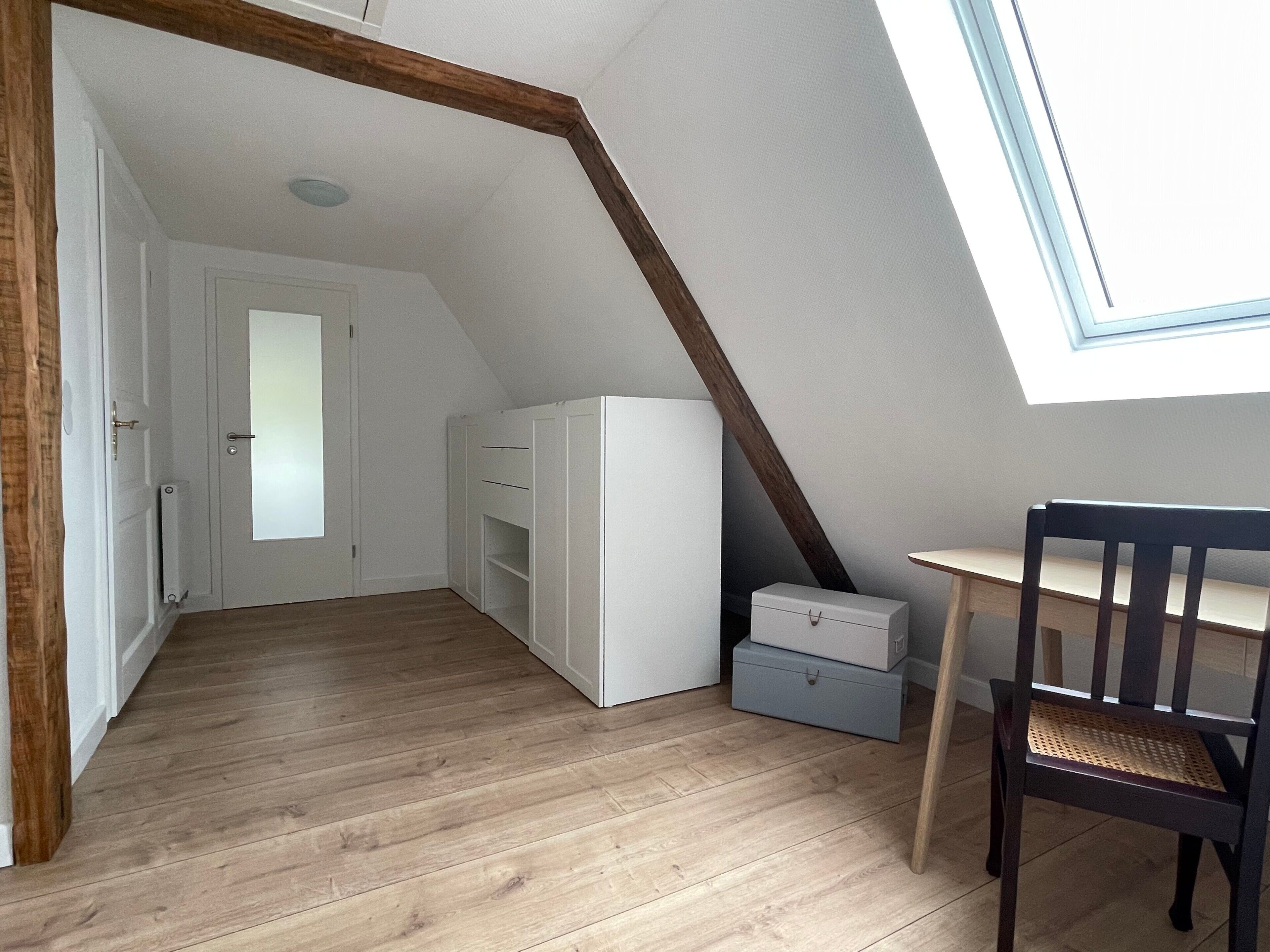 Een kamer met een schuine kap, houten balken, een witte garderobe, een tafeltje en een stoel.