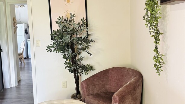 A corner room with a tufted ottoman, a brown armchair, a framed artwork, a potted plant, and a built-in shelf with a hanging plant. - Soleil Salem- extended stays, free laundry, garden (Salem)