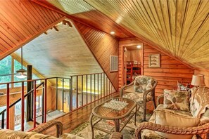A cozy wooden cabin interior with a high ceiling, a staircase leading to a loft area, wicker furniture, and a chessboard.