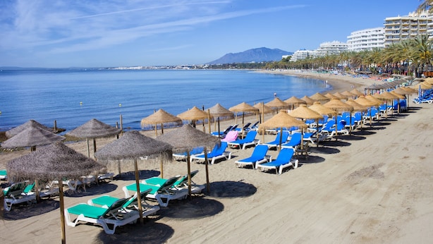 A beach with rows of blue lounge chairs and thatched umbrellas, a clear blue sky, and a mountain in the background. - Sunset Seaview Corner With Pool, Estepona, Spain (Estepona)