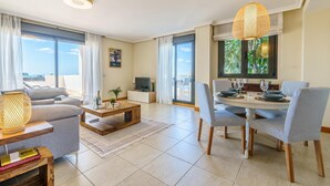 A modern living room with a sofa, coffee table, dining table with chairs, and a TV. - Sunset Seaview Corner With Pool, Estepona, Spain (Estepona)