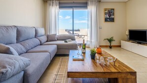 A modern living room with a grey sofa, a wooden coffee table with a bottle, glasses, and a book, and a TV on a white cabinet. - Sunset Seaview Corner With Pool, Estepona, Spain (Estepona)