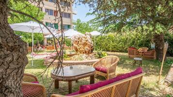 An outdoor seating area with wicker furniture and umbrellas, surrounded by trees and a building in the background.