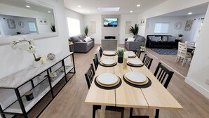 Een moderne eethoek met een houten tafel voor zes personen, omringd door stoelen. Achtergrond is een bank en een televisie.