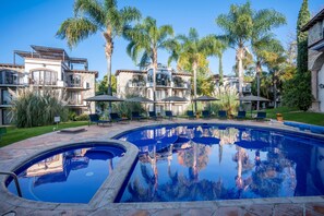 Una piscina con sillones de descanso y sombrillas, rodeada de palmeras y un edificio de varios pisos con balcones.