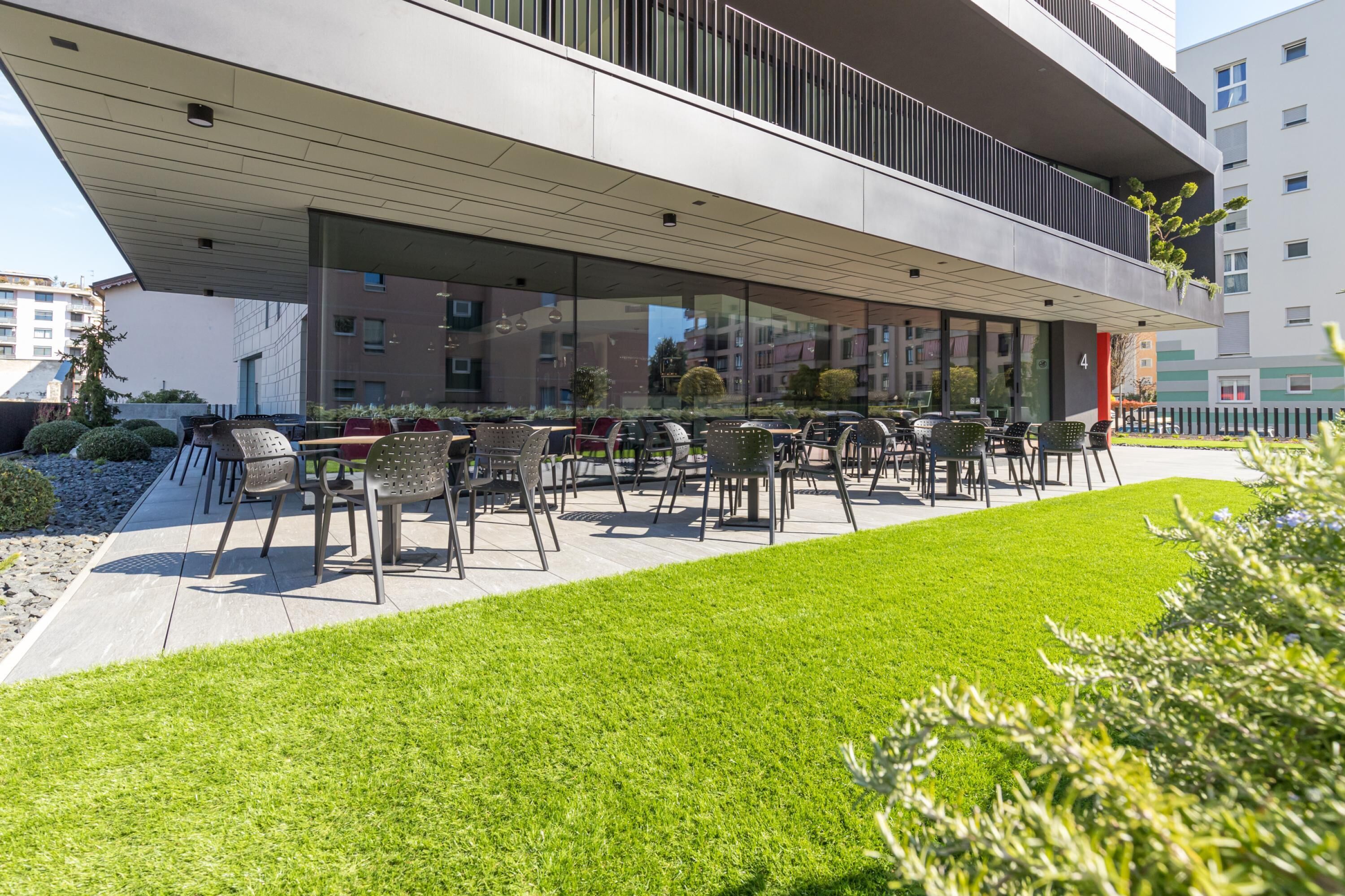 Un espace extérieur moderne aménagé avec des chaises et des tables, entouré d’une pelouse bien entretenue et d’un bâtiment doté de grandes fenêtres.