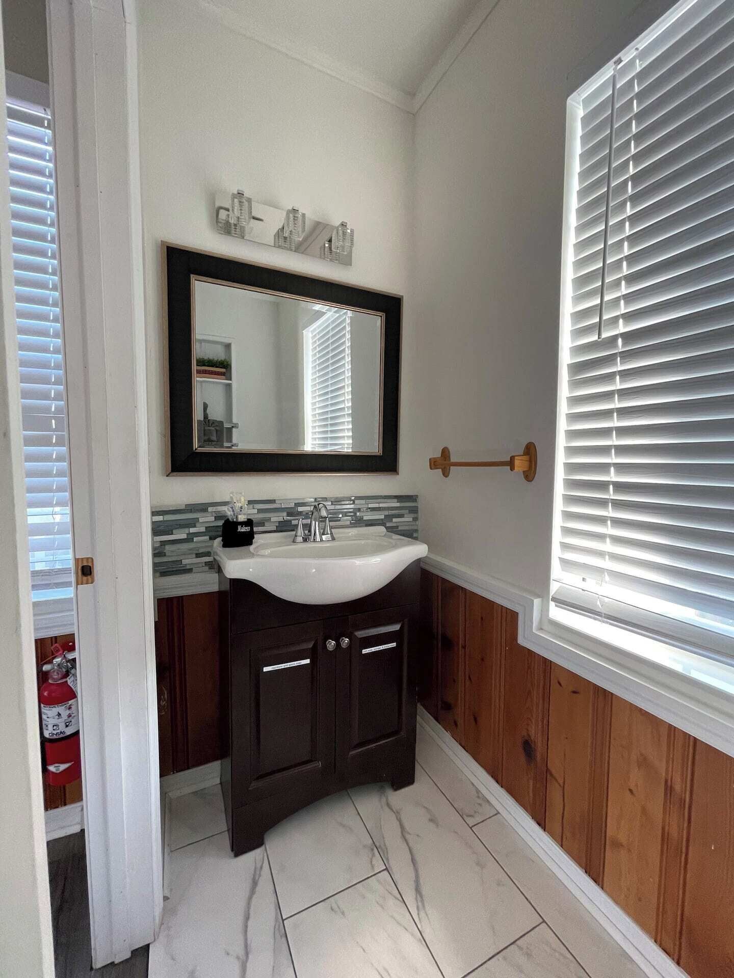 A bathroom with a white sink, dark wooden cabinets, a large mirror, and a window with blinds.