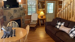 A cozy cabin interior with a stone fireplace, wooden walls, a wooden dining table with checkered placemats, a brown sofa with checkered cushions, and a staircase leading to an upper level. - Beautiful Authentic Log Cabin on Cosby Creek (Cosby)