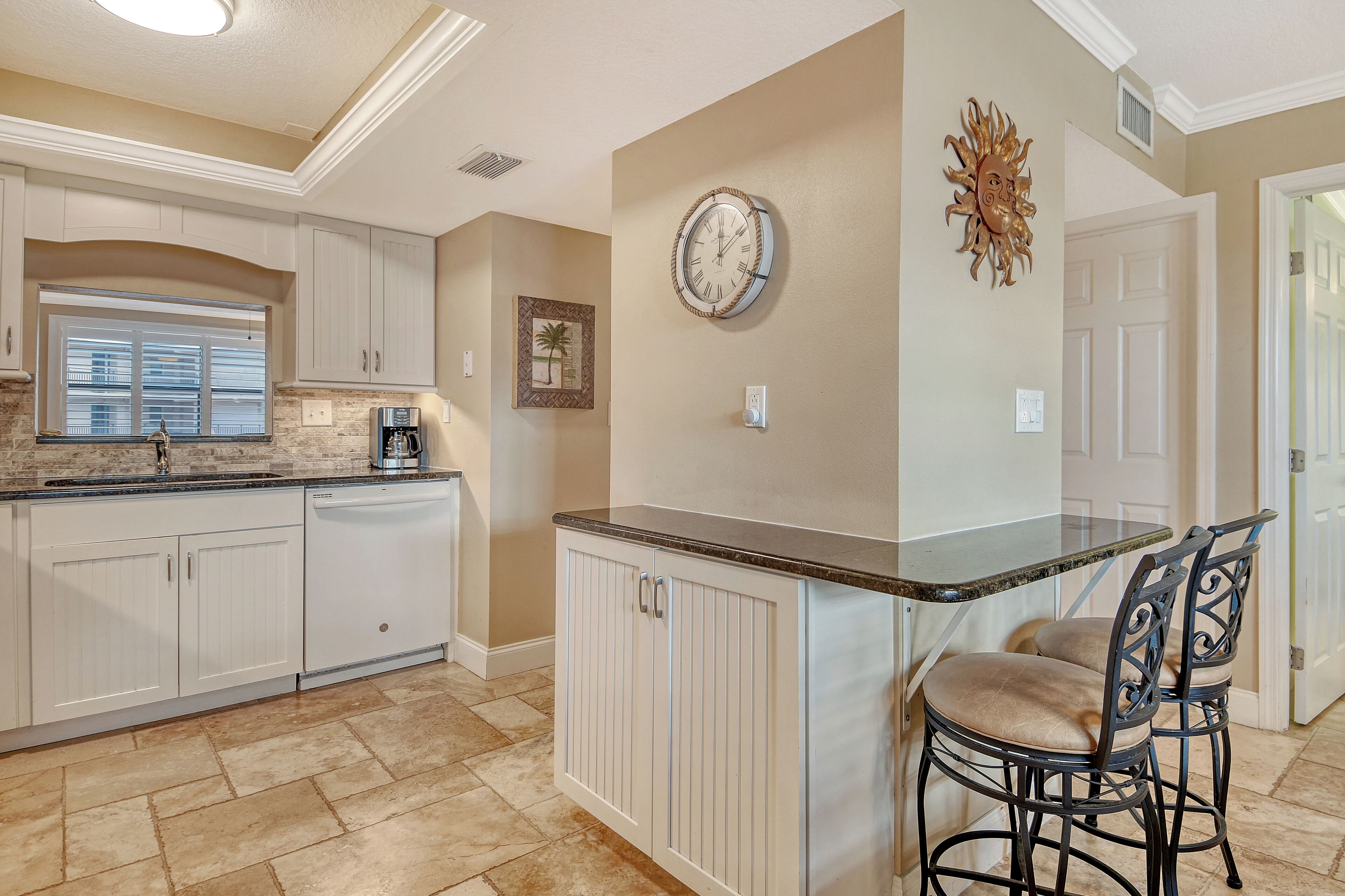 A kitchen with white cabinetry, a granite countertop, a built-in island with bar stools, a wall clock, and a decorative sun art piece.