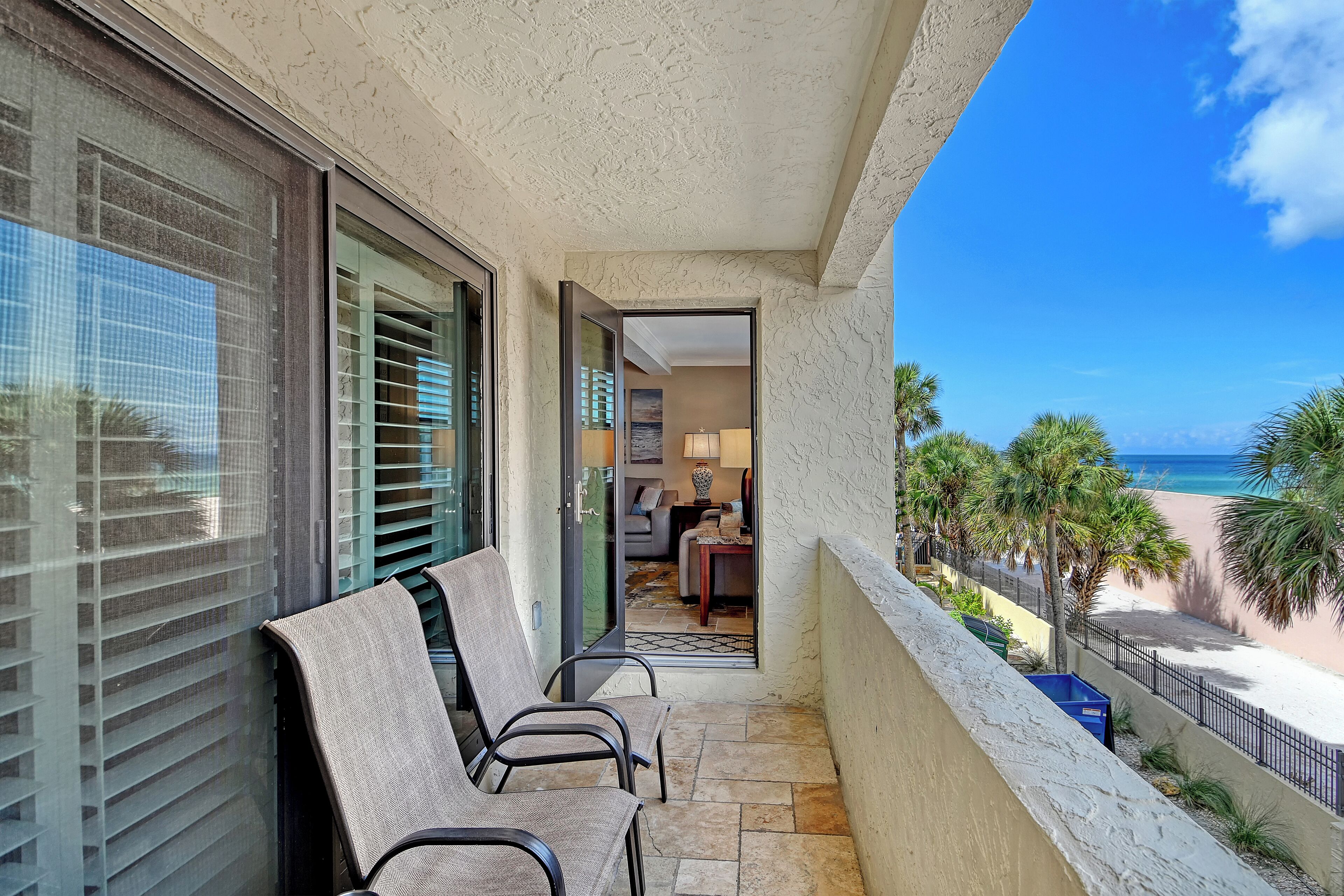 A balcony with two chairs, a view of the beach, and a door leading to an interior space.