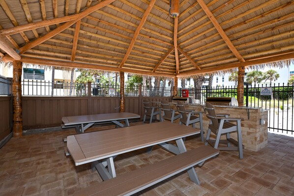 An outdoor patio with wooden tables and benches, a stone barbecue area, and a view of a fenced property with palm trees.