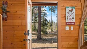 A wooden cabin entrance with a door leading to a forested area. A welcome sign with a moose and ski symbols is visible on the wall. - Timber Cabin in the woods (Cle Elum)