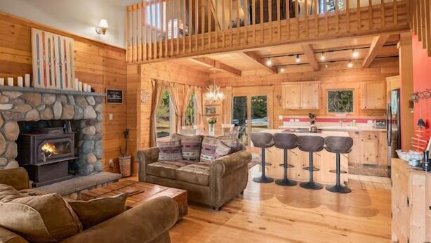 A rustic cabin interior with a stone fireplace, wooden floors, a lofted seating area, and a kitchen with wooden cabinets. - Timber Cabin in the woods (Cle Elum)