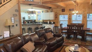 Un intérieur de chalet chaleureux avec un canapé en cuir, une table à manger avec des chaises, un coin cuisine et une vue sur l’extérieur.