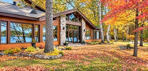 Une maison moderne dotée de grandes fenêtres, d’une cheminée en pierre et d’une cour bien entretenue parsemée de feuilles d’automne.