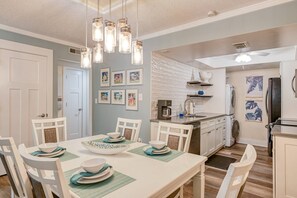Un coin repas avec une table blanche dressée pour quatre personnes, une cuisine avec des meubles blancs et une machine à café, et un couloir orné de tableaux encadrés.