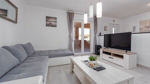 A modern living room with a grey sectional sofa, a white coffee table with decorative items, a flat-screen TV on a wooden stand, and a sliding glass door leading to an outdoor area. - Villa Jenny (Policnik)