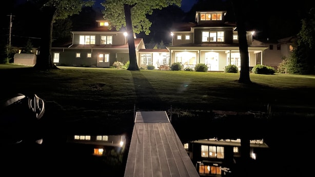 A two-story house with a well-lit front porch and a dock extending into a body of water.