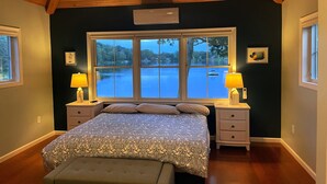 A bedroom with a large window overlooking a lake, a bed with patterned bedding, two bedside tables with lamps, and a wooden ceiling.