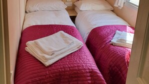 Quarto pequeno com duas camas, cada uma com colcha rosa e travesseiros brancos. Há armários de madeira sobre as camas e uma janela com cortinas.